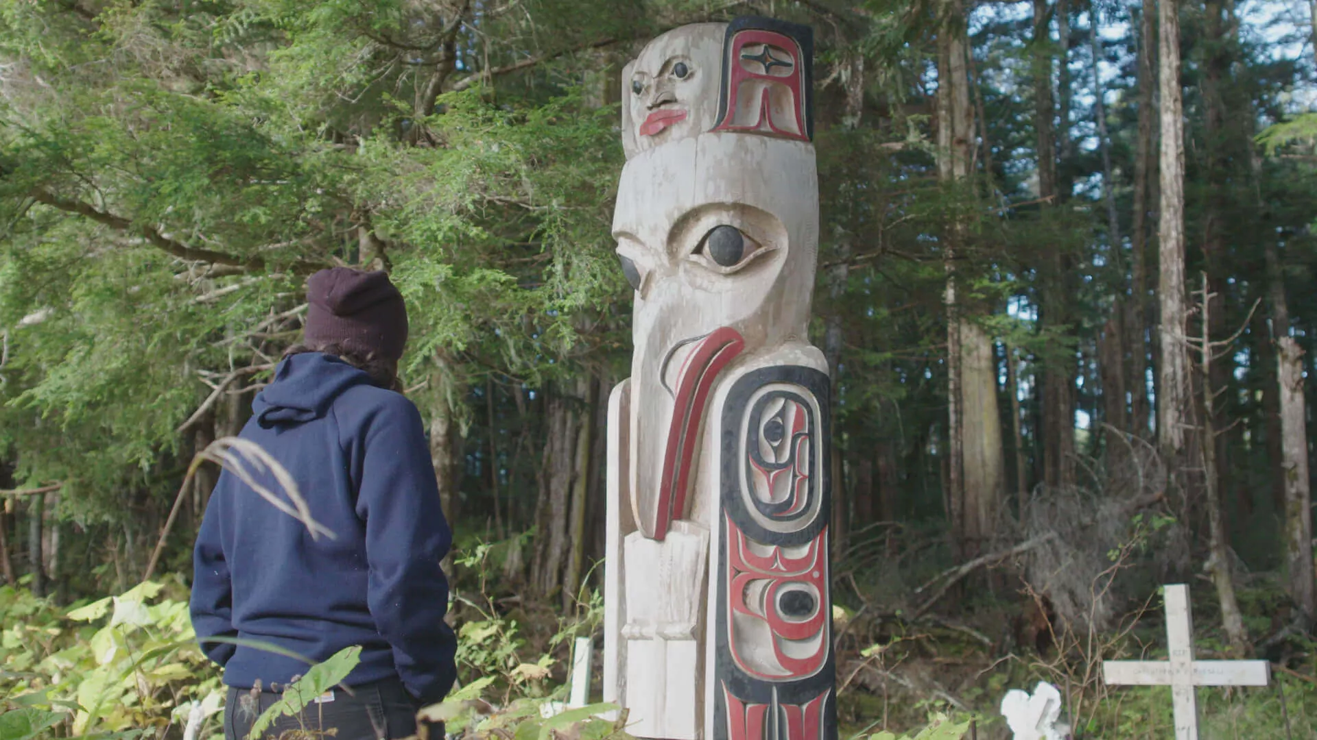 A lone traveller examines a large totem statue in a forest, painted in white, red, and black. (Skindigenous Season 3)
